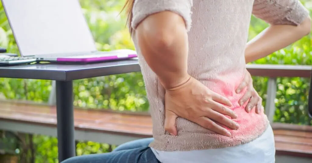 Mujer sentada a la mesa mientras se sujeta la parte baja de la espalda