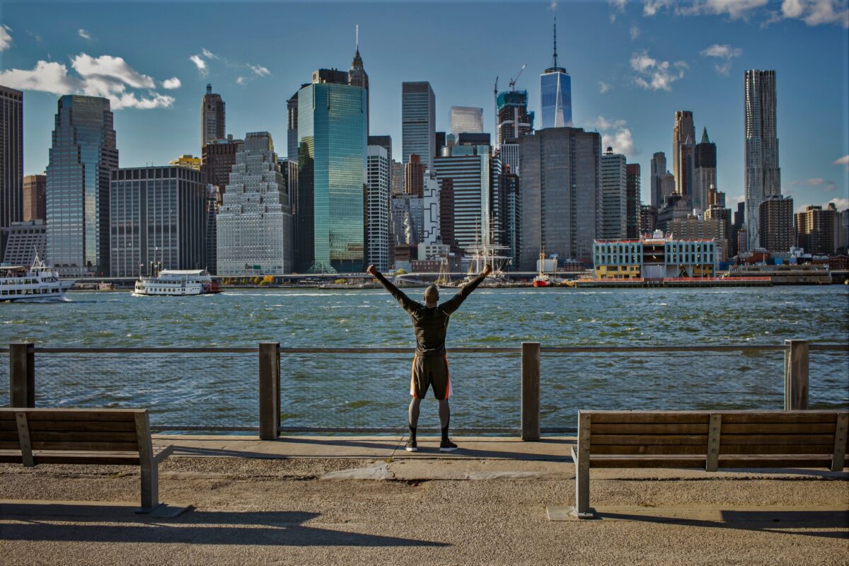 Vista trasera de un joven atleta emocionado de pie en el paseo marítimo con las manos levantadas contra el horizonte del Bajo Manhattan
